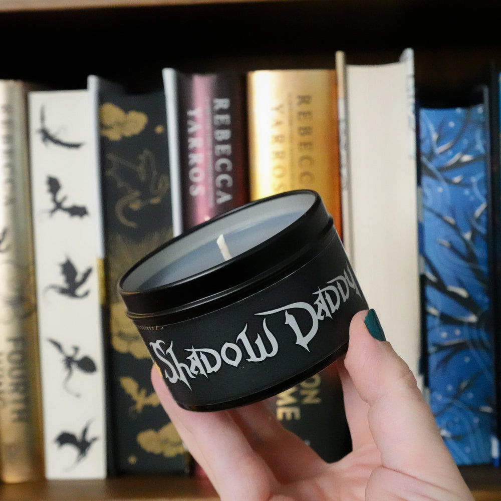 A black soy wax candle with a label that reads 'Shadow Daddy'. The candle is placed on a surface with books and a decorative item.