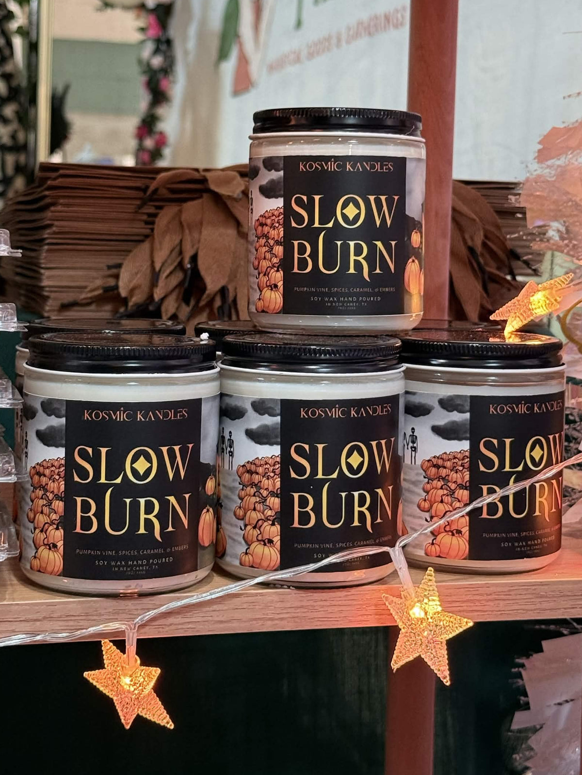 Several jars of Kosmic Kandles Slow Burn  candles are displayed on wooden shelves decorated with warm star-shaped string lights.
