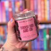 Hand holding a 'Kosmic Kanes' candle labeled 'Touch Her and Die' in front of a bookshelf.