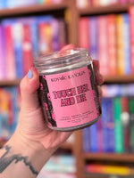 Hand holding a 'Kosmic Kanes' candle labeled 'Touch Her and Die' in front of a bookshelf.
