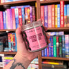 Hand holding a 'Kosmic Kandles' candle labeled 'Touch Her and Die' in front of a bookshelf.