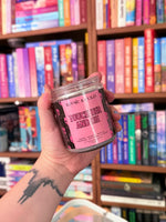 Hand holding a 'Kosmic Kandles' candle labeled 'Touch Her and Die' in front of a bookshelf.