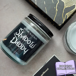 Candle jar labeled 'Shadow Daddy' from Kosmic Kandles on a surface with book-themed packaging.